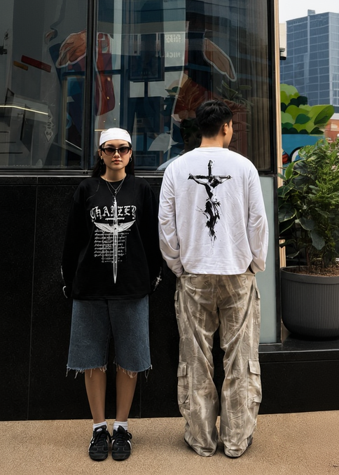 Two models standing in front of a glass storefront with reflections and colorful mural art in the background. One model is wearing a black graphic sweatshirt, denim shorts, and a white cap, while the other is wearing a white graphic sweatshirt and beige cargo pants.